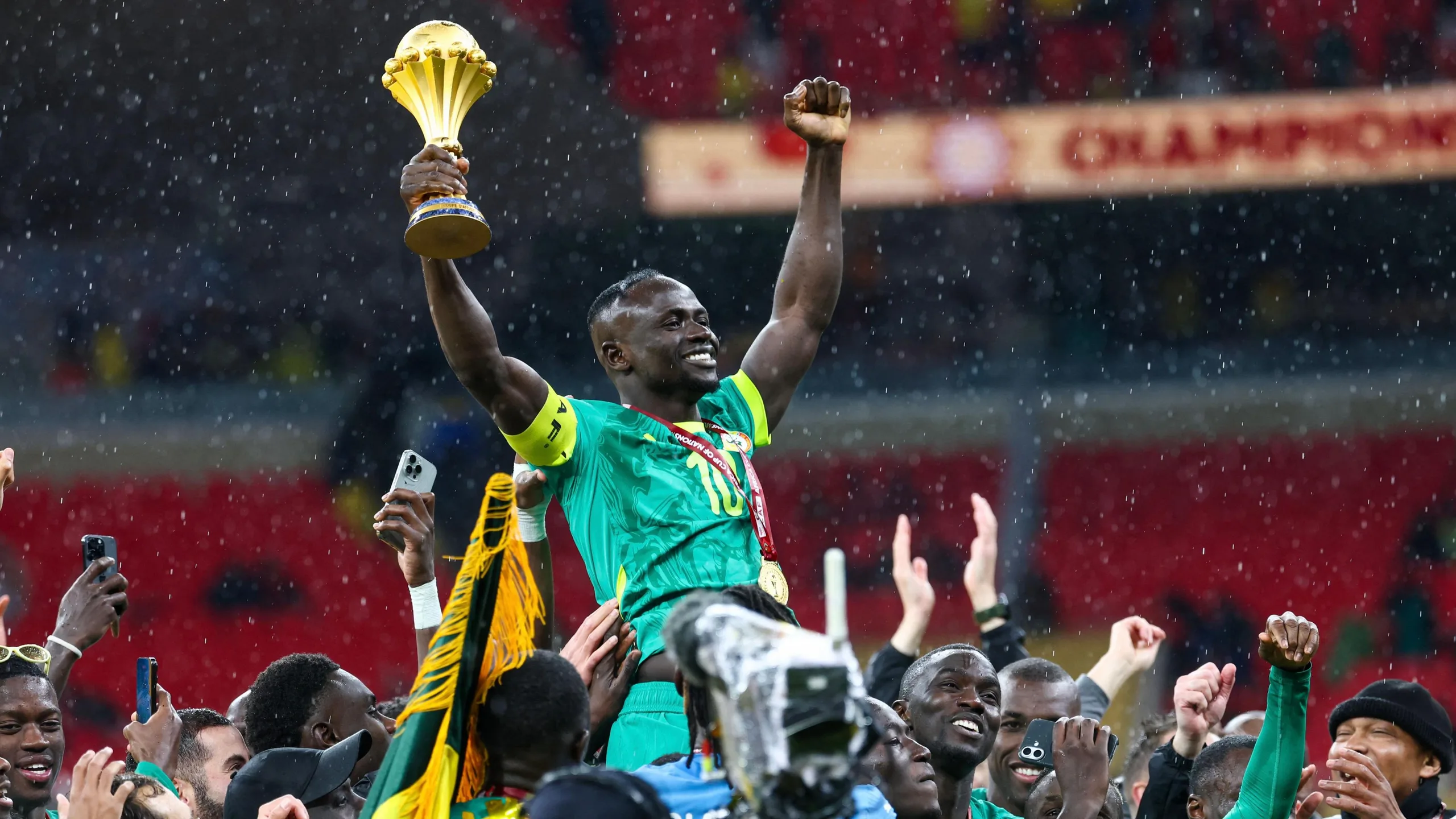Sadio Mane, wearing green Senegal kit and with a golden medal around his neck on a red ribbon, is raised on the shoulders of his team-mates after the final of the 2025 Afcon. As drops of rain fall, Mane raises both arms in the air, clutching the Afcon trophy in his right hand. The heads of the group gathered around him can be seen, with a mix of hands also raised, some clapping, others filming on phones, while a TV camera with a rainproof cover is also visible in the throng