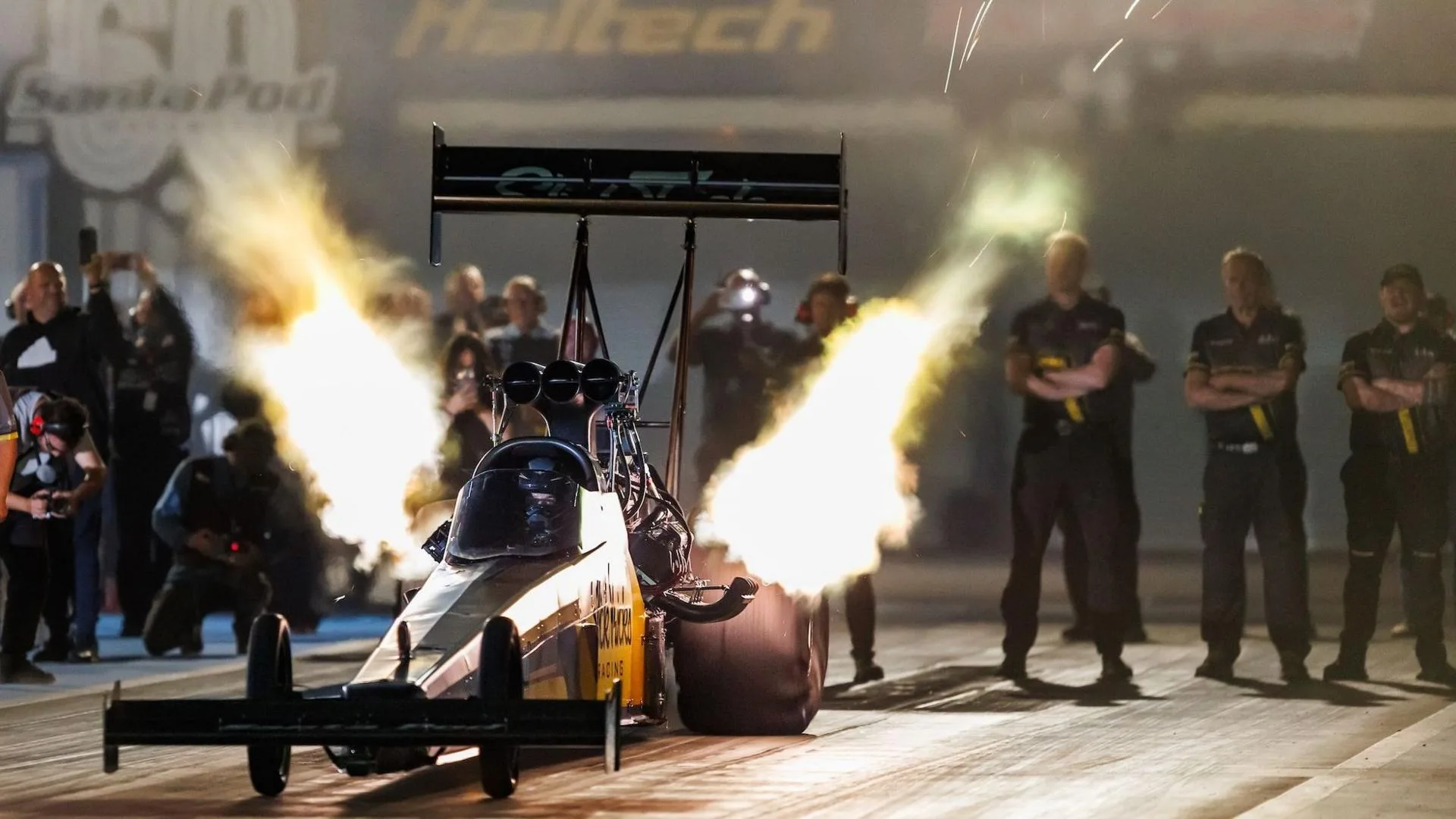 Susanne Callin sets of in her Top Fuel dragster at Santa Pod near Northampton, in September 2026. Her car is pictured in low light, with large blasts of flame on either side above the large back wheels. Her team stand behind as the car leaves the starting line.
