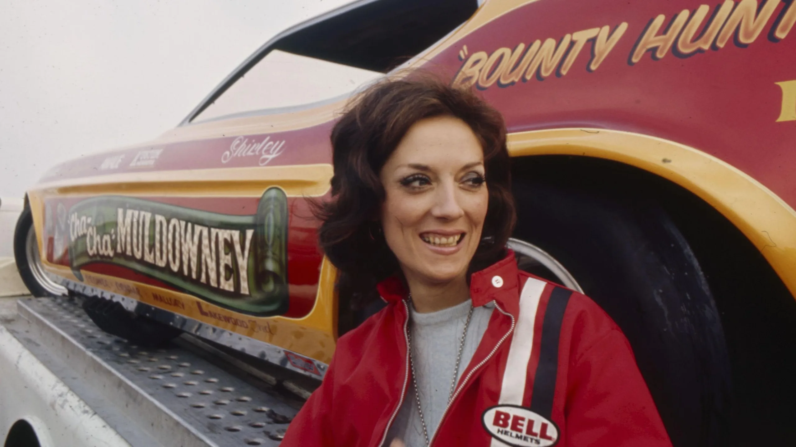 Drag racing trailblazer Shirley Muldowney pictured at the National Hot Rod Association (NHRA) Winternationals at Pomona Raceway in 1970. She is wearing a red jacket and smiling, in front of her car, which is red with "Bounty Hunter" and her name painted on it in gold-coloured writing.