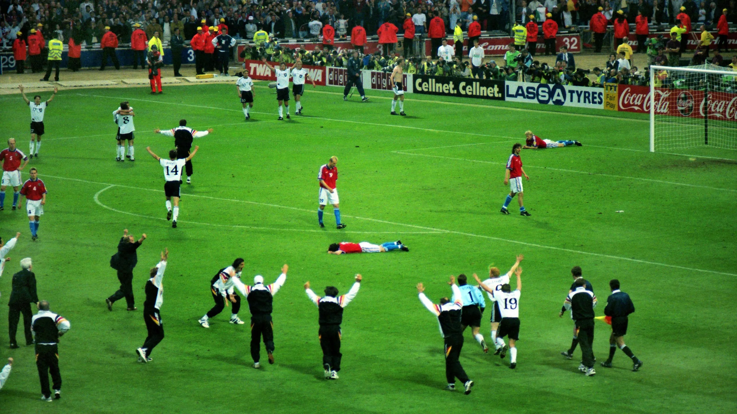Germany players, in black shirts and black shorts, celebrate their Euro '96 win after a golden goal in extra time as Czech players in red shirts look distraught, with one lying face down on the pitch