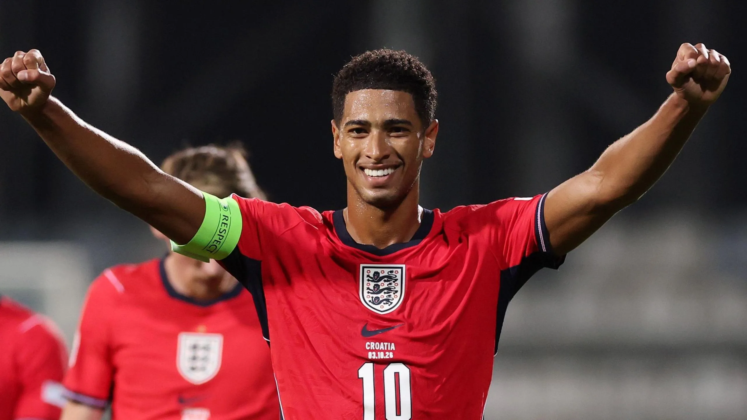 A smiling Jude Bellingham, in red kit, celebrates during England's win over Croatia with both arms stretched out wide to head height