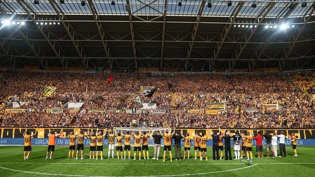 Dynamo Dresden players stand in front of a large, seemingly packed, stand of their own supporters, raising their arms to the fans