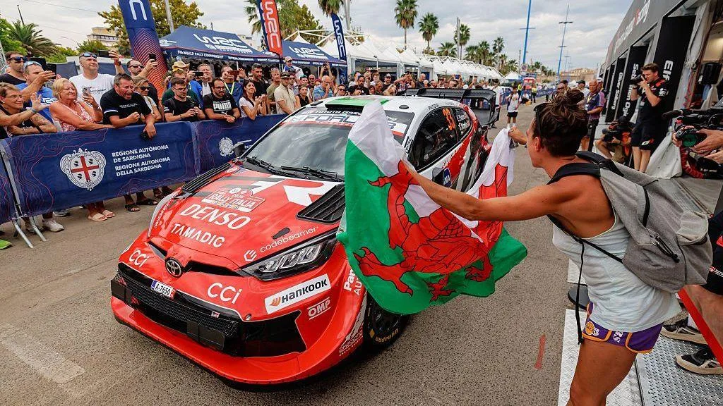 A fan waves a Welsh flag in front of a rally car while fans behind cheer