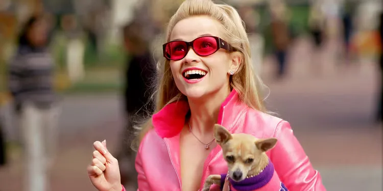 Reese Witherspoon as Elle Woods smilingly widely in a pink outfit holding a small dog in Legally Blonde.