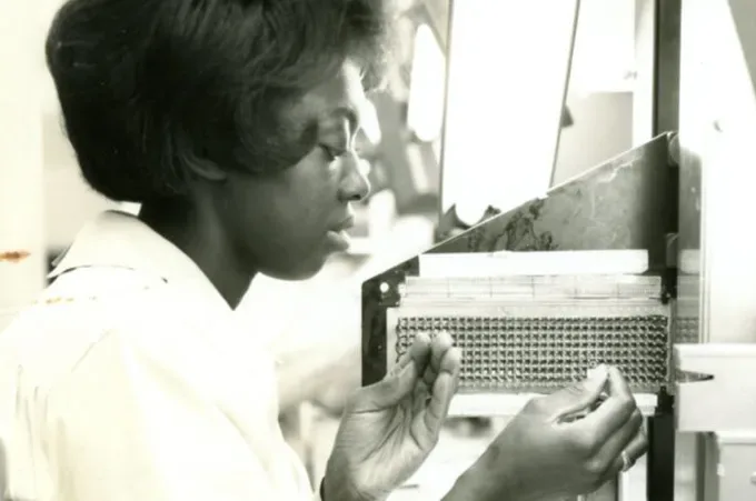 an old, black-and-white photo of a woman threading metal wire through small holes in a machine