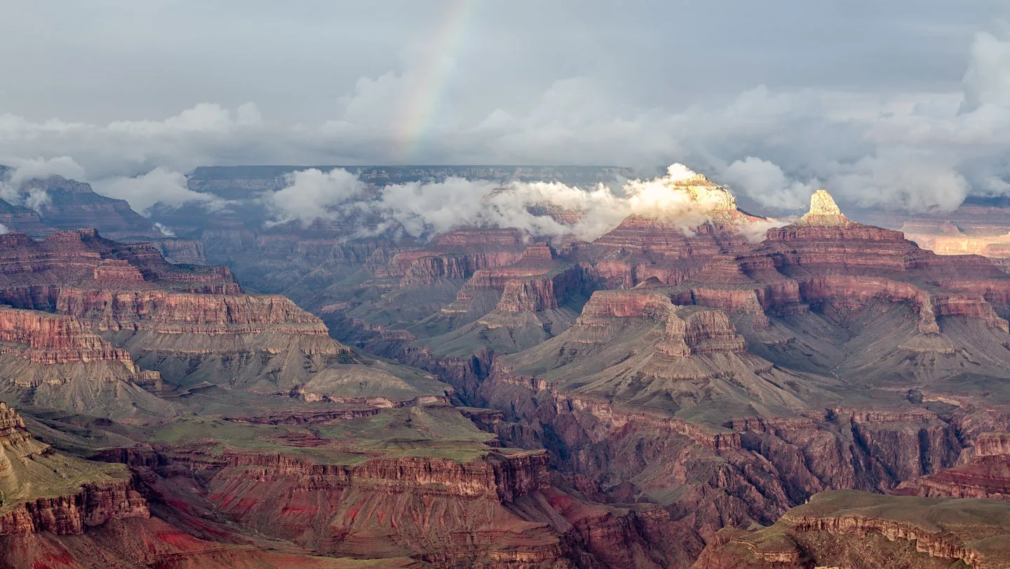 A faint rainbow arcs over the exposed rock layers of the Grand Canyon.