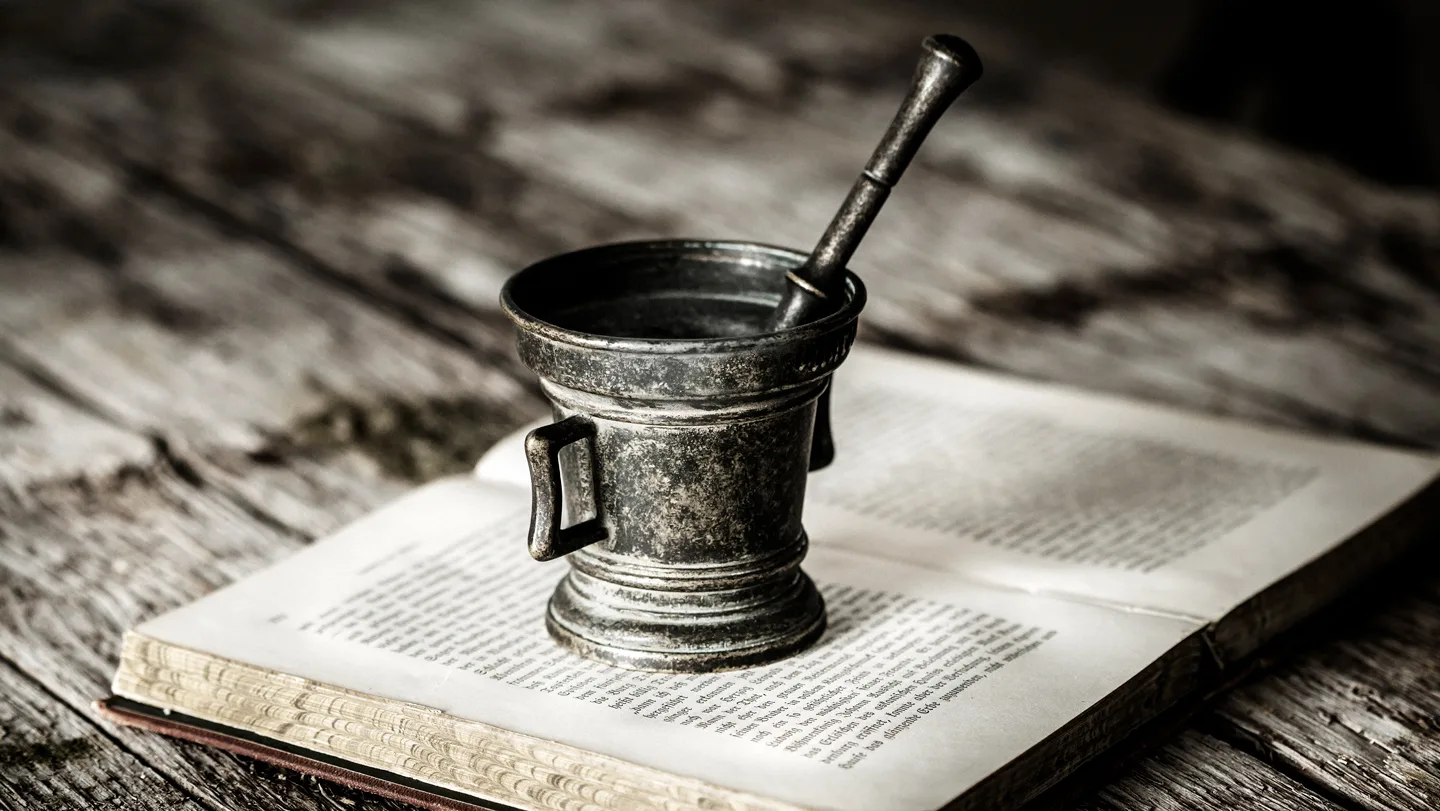 A tarnished metal cup with a stirrer sits on top of an open, dusty-looking book on a wood table.